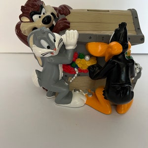 May include: A ceramic piggy bank shaped like a treasure chest with Bugs Bunny, Daffy Duck, and Tasmanian Devil characters. The chest is open and the characters are looking inside. The chest is decorated with gold coins and jewels.
