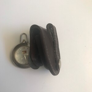 May include: A silver pocket watch with a white face and black hands. The watch is in a brown leather case with a silver button.