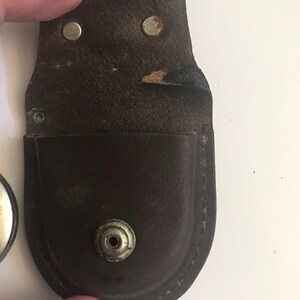 May include: A brown leather case with a snap closure and a metal button. The case is designed to hold a small, round object.