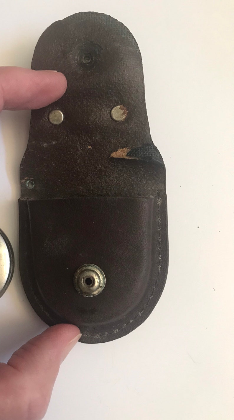 May include: A brown leather case with a snap closure and a metal button. The case is designed to hold a small, round object.