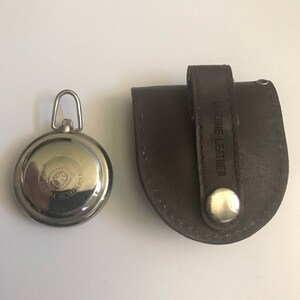 May include: A silver pocket watch with a black face and a leather carrying case. The case has the words "Genuine Leather" embossed on it.