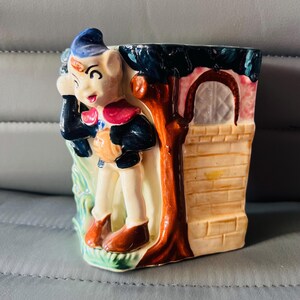 May include: A ceramic planter shaped like a small house with a whimsical elf figure leaning against a tree. The elf is wearing a blue hat, red and blue shirt, and brown shoes. The house has a brown door and a brick wall.