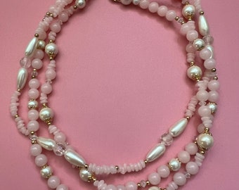 May include: A three-strand necklace with a pink background. The necklace features a mix of pink beads, pearls, and gold-colored accents. The strands are connected with a gold-colored clasp.
