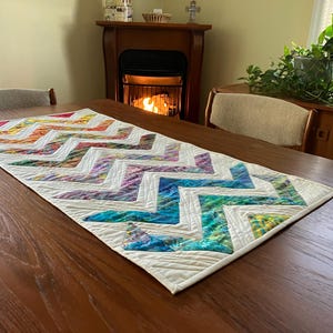 May include: A white table runner with a chevron pattern in shades of rainbow colors. The runner is on a wooden table with a fireplace in the background.