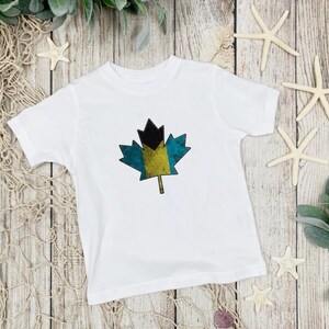 May include: White t-shirt with a distressed, blue, yellow, and black maple leaf design. The maple leaf is a stylized version of the flag of the Bahamas.