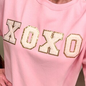 May include: Pink sweatshirt with a gold and white XOXO graphic.