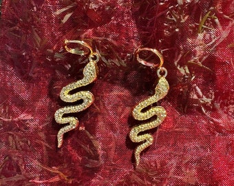 May include: A pair of gold-colored snake earrings with sparkling embellishments. The earrings are displayed on a red, sheer fabric, creating a striking contrast. The snake design is detailed and elegant, perfect for adding a touch of glamour.