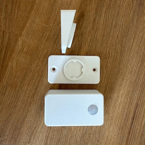 Può includere: Un dispositivo sensore di movimento rettangolare bianco con una lente circolare sulla parte anteriore, una staffa di montaggio separata con un'indentazione circolare centrale e un piccolo componente in plastica bianca a forma di cuneo.
