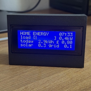 May include: A black rectangular energy monitor with a bright blue backlit LCD screen displaying text including HOME ENERGY, 07:33, load 0.4kW, today 2.9kWh, 0.80, solar 0.3, and grid 0.1. The device is housed in a matte black 3D printed enclosure.
