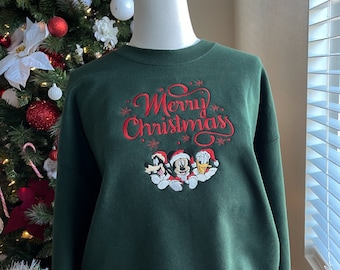 May include: A dark green sweatshirt with a red embroidered design that says "Merry Christmas" with Mickey Mouse, Donald Duck, and Goofy wearing Santa hats.
