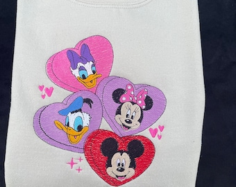 May include: A light beige sweatshirt with a graphic of Mickey Mouse, Minnie Mouse, Donald Duck, and Daisy Duck in heart-shaped candy boxes. The hearts are pink, purple, and red.