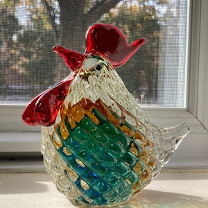 May include: A decorative glass rooster figurine with a red comb and wattle. The body features a textured, quilted design with colors of green, gold, and blue. The rooster is positioned on a windowsill with a tree visible in the background.