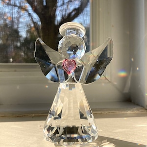 May include: A clear crystal angel figurine with a faceted design. The angel has wings, a halo, and a pink heart-shaped gem in the center. The figurine is positioned on a white surface, with sunlight creating a rainbow effect.