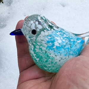 May include: A small glass bird figurine with a blue beak and white and blue speckled body. The bird is sitting on a hand.