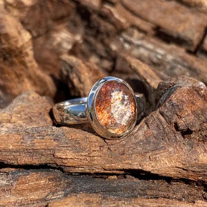 May include: A silver ring with a round, iridescent sunstone gemstone. The stone has a mix of orange, red, and gold colors.