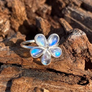 Op de afbeelding: Een zilveren ring met een bloemdesign. De ring heeft vijf bloembladen, elk gemaakt van een iriserende steen met blauwe en witte tinten. De ring ligt op een getextureerd, bruin houten oppervlak.