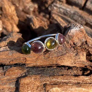 May include: A silver ring featuring five colorful gemstones. The gemstones are a mix of green, purple, and yellow, set in individual silver bezels. The ring is displayed on a textured, brown wooden surface.