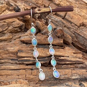 Può includere: Orecchini pendenti in argento con opali ovali. Ogni orecchino presenta sei opali nei toni del blu, verde e rosa. Gli opali sono collegati da piccoli anelli d'argento, creando un design fluido ed elegante.
