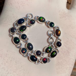 Peut inclure: Un bracelet en argent orné d'opales noires ovales avec des éclats verts, rouges et oranges. Les opales sont serties dans une chaîne, créant un bijou saisissant.