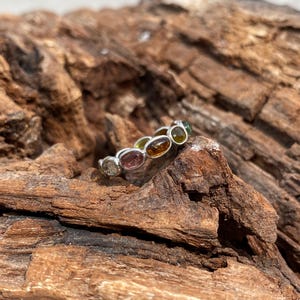 May include: A silver ring with a row of oval-shaped gemstones in various colors, including pink, green, yellow, and brown. The ring is set on a piece of weathered wood.