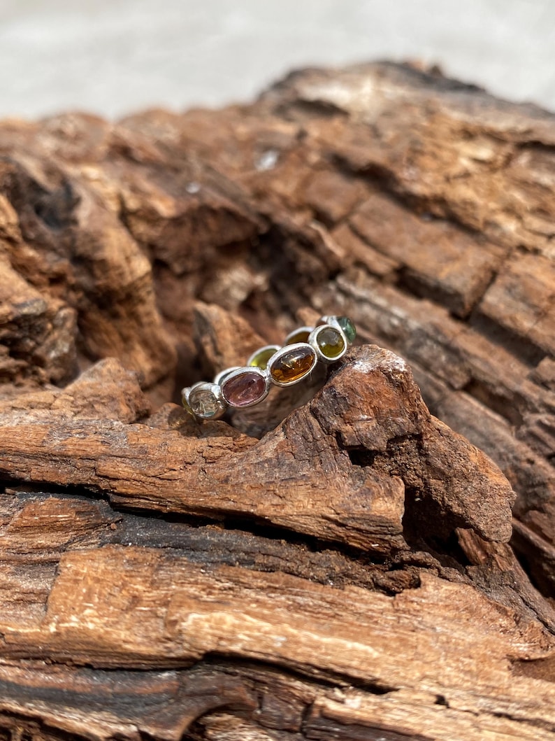May include: A silver ring with a row of oval-shaped gemstones in various colors, including pink, green, yellow, and brown. The ring is set on a piece of weathered wood.