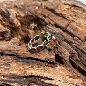 May include: A silver ring with a band of oval-shaped gemstones in shades of green, brown, and pink.