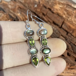 May include: Silver dangle earrings with green and brown gemstones. The earrings have a teardrop shape with a silver hook.