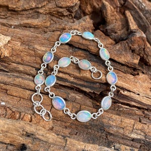 Può includere: Un bracciale in argento con opali ovali. Gli opali mostrano colori come blu, rosa e bianco. Il bracciale è posto su uno sfondo di legno strutturato, evidenziando il design e l'artigianato del gioiello.