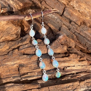 May include: A pair of silver dangle earrings featuring a series of oval and teardrop-shaped opals. The opals have a milky white base with flashes of blue and green. The earrings are suspended from a silver hook.