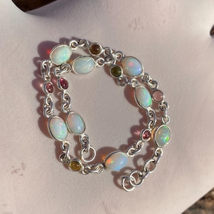 May include: A silver bracelet with oval opals and smaller gemstones in pink, brown, and green. The chain link bracelet is displayed on a light pink surface. The gemstones are set in a silver chain.