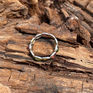 May include: A silver ring with a row of oval-shaped gemstones in various colors, including green, red, and yellow. The ring is on a piece of weathered wood.