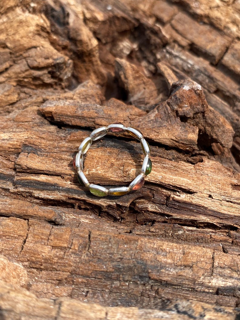 May include: A silver ring with a row of oval-shaped gemstones in various colors, including green, red, and yellow. The ring is on a piece of weathered wood.