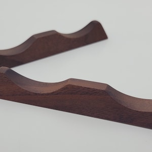 May include: Two wooden wine bottle cradles. Each cradle is a dark brown color, with a curved design to hold a bottle. The cradles are made of solid wood and have a smooth finish. They are designed to hold a standard size wine bottle.