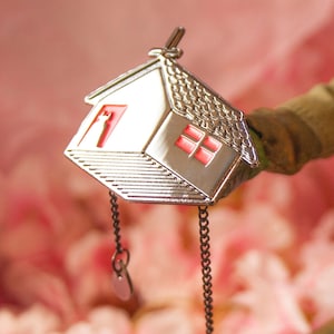 Può includere: Un ciondolo d'argento a forma di casa con una finestra rossa e una catena attaccata. Il ciondolo è appeso su uno sfondo rosa sfocato.