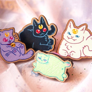 Könnte beinhalten: Vier hölzerne Katzennadeln mit verschiedenen Farben und Designs. Die Katzen sitzen alle in einer sitzenden Position mit einem Halbmond auf der Stirn. Die Nadeln sind in Lila, Schwarz, Weiß und Grün bemalt.