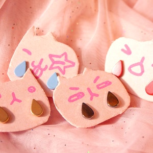 Könnte beinhalten: Vier rosafarbene, katzenförmige Ohrringe aus Ton mit verschiedenen Gesichtsausdrücken und tropfenförmigen goldenen, braunen und blauen Verzierungen.