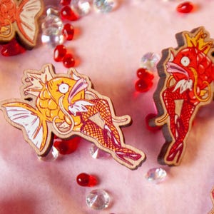 Könnte beinhalten: Zwei Holz-Anstecknadeln mit Cartoon-Fischen mit menschlichen Beinen, die Netzstrümpfe tragen. Ein Fisch ist rot und der andere ist gelb. Beide Fische haben Kronen auf dem Kopf.