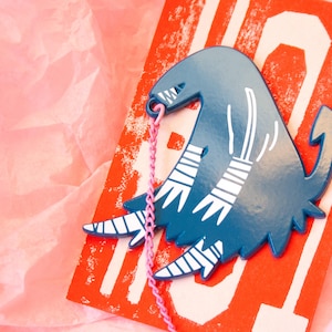 Può includere: Una creatura in ceramica blu e bianca con uno spago rosa attaccato. La creatura ha una lunga coda e strisce bianche sulle gambe. La creatura è seduta su uno sfondo rosso e bianco.