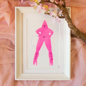 Puede incluir: Impresión enmarcada de una figura rosa con una pose segura, con botas de tacón alto. La obra de arte está enmarcada en un marco blanco. El fondo incluye tela rosa y una rama con flores rosas, añadiendo un toque decorativo.