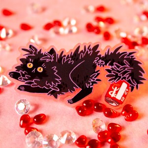 Könnte beinhalten: Eine schwarze Katze mit einem flauschigen Schwanz hält eine rote Dose mit weißem Text. Die Katze liegt auf einer rosa Oberfläche mit roten und klaren Edelsteinen, die um sie herum verstreut sind.