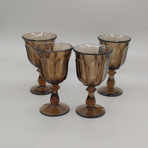 May include: Four brown glass goblets with a fluted design. The goblets are arranged in a semi-circle on a white surface.