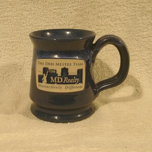 May include: A blue ceramic coffee mug with a gold logo that reads "The Debi Mestre Team STAR MD Realty Distinctively Different".