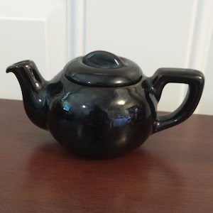 May include: A small, black ceramic teapot with a shiny glaze. The teapot has a rounded body and a short spout.