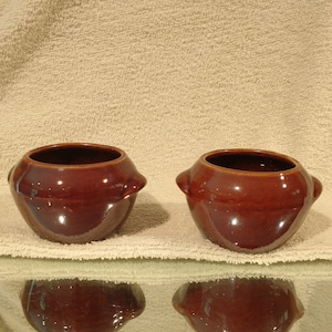 May include: Two brown ceramic bowls with rounded edges and a glazed finish. The bowls are sitting on a light brown fuzzy surface.