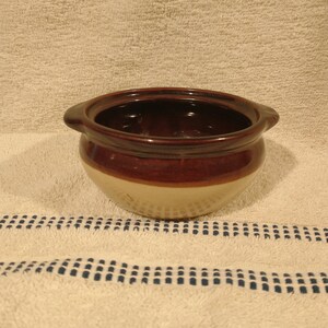 May include: A brown and tan ceramic bowl with a rounded shape and two handles. The bowl is glazed and has a rustic look.