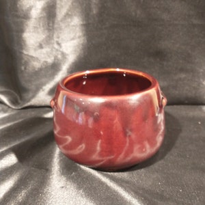 May include: A small, dark brown ceramic pot with two small handles on the sides. The pot is glazed and has a smooth, rounded shape.
