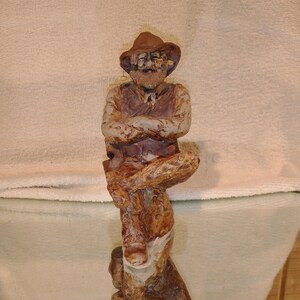 May include: A ceramic figurine of a man wearing a brown hat and a vest. He is sitting with his arms crossed and has a pipe in his mouth. The figurine is painted in shades of brown, white, and black.