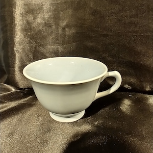 May include: A light blue teacup with a handle and a small base. The cup is sitting on a dark brown surface.