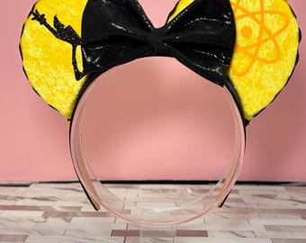 Op de afbeelding: Gele fluwelen Minnie Mouse oren met een zwarte strik en een silhouet van een personage dat een toverstaf vasthoudt op het ene oor en een atoomsymbool op het andere oor.