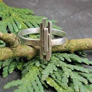 Puede incluir: Un anillo plateado con un diseño de barras verticales. El anillo se encuentra sobre un fondo de follaje verde y una superficie gris oscuro. El diseño del anillo presenta varias barras rectangulares de diferentes longitudes.
